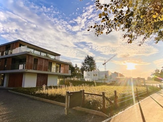 A modern building with large glass windows and wooden accents stands near a construction site. The sun is setting, casting a warm glow over the scene. The sky is filled with scattered clouds, and a construction crane is visible in the background. Trees and a fence line the area, contributing to a serene and peaceful atmosphere.