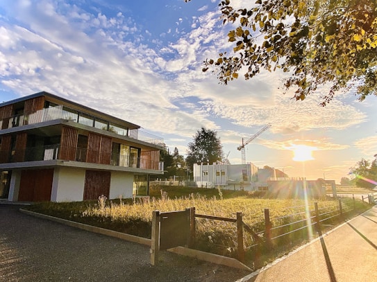 A modern building with large glass windows and wooden accents stands near a construction site. The sun is setting, casting a warm glow over the scene. The sky is filled with scattered clouds, and a construction crane is visible in the background. Trees and a fence line the area, contributing to a serene and peaceful atmosphere.