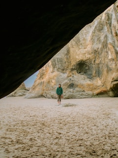 A person stands barefoot on a sandy beach, surrounded by large rocky formations. The individual wears a green jacket and carries a pair of shoes. The lighting is natural, and there is an overcast sky visible above the rocks.