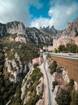 A scenic view of rocky mountains with lush greenery and architectural structures nestled among the cliffs. A funicular railway track runs through the terrain, leading towards the building with a red roof. The sky is partly cloudy, enhancing the majestic and tranquil atmosphere.