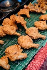 Pieces of crispy, golden-brown fried chicken are arranged on a metal cooling rack. The chicken is placed on top of a green banana leaf, which rests on a red and white polka dot cloth.
