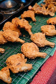 Pieces of crispy, golden-brown fried chicken are arranged on a metal cooling rack. The chicken is placed on top of a green banana leaf, which rests on a red and white polka dot cloth.