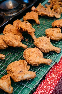 Pieces of crispy, golden-brown fried chicken are arranged on a metal cooling rack. The chicken is placed on top of a green banana leaf, which rests on a red and white polka dot cloth.