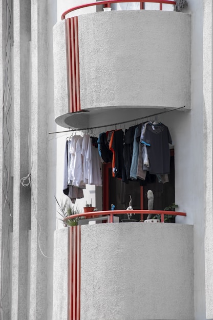 A small balcony with a red railing, featuring clothes hanging on a line to dry. The balcony has a rounded, textured concrete wall with vertical red stripes. Potted plants are visible on the balcony ledge, adding greenery to the scene. The background consists of a light-colored wall with vertical shadows from possibly architectural elements.