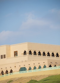 A large, beige building with intricate arched windows and a decorative trim along the roofline. The structure is situated on a hill, surrounded by green grass, and set against a clear blue sky.