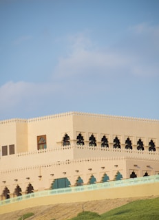 A large, beige building with intricate arched windows and a decorative trim along the roofline. The structure is situated on a hill, surrounded by green grass, and set against a clear blue sky.