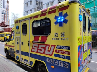 A yellow ambulance with blue and red markings is parked on a city street. The vehicle has various inscriptions, such as 'AMBULANCE' and 'SPECIAL SUPPORT UNIT', along with emergency symbols and Chinese characters. Blue emergency lights are visible on the sides. Nearby, buildings and another vehicle can be seen, including a crane in the background.
