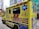 A yellow ambulance with blue and red markings is parked on a city street. The vehicle has various inscriptions, such as 'AMBULANCE' and 'SPECIAL SUPPORT UNIT', along with emergency symbols and Chinese characters. Blue emergency lights are visible on the sides. Nearby, buildings and another vehicle can be seen, including a crane in the background.