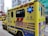A yellow ambulance with blue and red markings is parked on a city street. The vehicle has various inscriptions, such as 'AMBULANCE' and 'SPECIAL SUPPORT UNIT', along with emergency symbols and Chinese characters. Blue emergency lights are visible on the sides. Nearby, buildings and another vehicle can be seen, including a crane in the background.