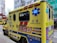 A yellow ambulance with blue and red markings is parked on a city street. The vehicle has various inscriptions, such as 'AMBULANCE' and 'SPECIAL SUPPORT UNIT', along with emergency symbols and Chinese characters. Blue emergency lights are visible on the sides. Nearby, buildings and another vehicle can be seen, including a crane in the background.