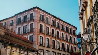 A multi-story brick building with numerous windows and black wrought-iron balconies stands prominently against a clear blue sky. It is flanked by other buildings, including a market sign reading 'Mercado de San Miguel'. The architecture is classic and elegant, with a warm afternoon light casting subtle shadows.