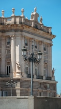 A historic building with neoclassical architecture features ornate details such as columns, statues, and balustrades. The bright blue sky serves as a backdrop, highlighting the intricate stone façade. A vintage streetlamp is prominently set in the foreground, adding to the classical aesthetic.