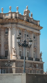 A historic building with neoclassical architecture features ornate details such as columns, statues, and balustrades. The bright blue sky serves as a backdrop, highlighting the intricate stone façade. A vintage streetlamp is prominently set in the foreground, adding to the classical aesthetic.