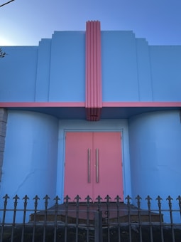 A building facade painted in pastel colors, featuring a prominent pink door flanked by blue walls. The architecture includes vertical grooves and a striking pink column running upwards. There is a decorative metal fence in front, adding an ornamental element.