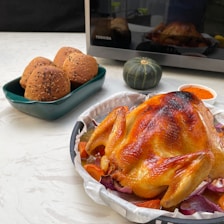 A beautifully roasted chicken is placed on a tray, surrounded by vegetables like carrots and red onions. Next to it is a rectangular dish containing three sesame-seeded brown bread rolls, a small green pumpkin, and a small bowl of orange spices. In the background, a microwave is visible.