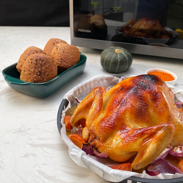 A beautifully roasted chicken is placed on a tray, surrounded by vegetables like carrots and red onions. Next to it is a rectangular dish containing three sesame-seeded brown bread rolls, a small green pumpkin, and a small bowl of orange spices. In the background, a microwave is visible.