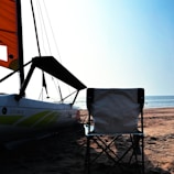 A beach scene with a catamaran visible in the foreground, featuring a brightly colored sail with orange and yellow patterns. A lone folding chair is placed on the sandy shore, facing the calm sea. The sky is clear with a light blue hue, indicating a serene and sunny day.