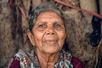 An elderly person with gray hair and a colorful scarf sits in front of a textured background. The expression is calm and composed, with a gentle gaze and slight smile.