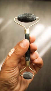 A hand is holding a jade roller, an object typically used for facial massage. The roller is made of a handle and two stone rollers, one larger at the top and one smaller at the bottom, both made from dark green jade.