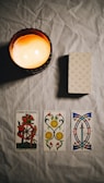 A dimly lit scene featuring a burning candle casting a warm glow over a table. On the table, there is a neatly stacked deck of patterned cards and three tarot cards laid out side by side. The cards have intricate designs, with the Wheel of Fortune card prominently displayed.