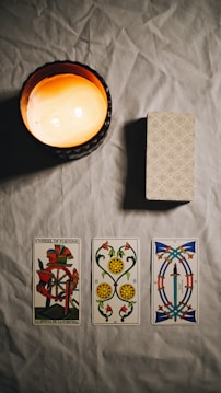 A dimly lit scene featuring a burning candle casting a warm glow over a table. On the table, there is a neatly stacked deck of patterned cards and three tarot cards laid out side by side. The cards have intricate designs, with the Wheel of Fortune card prominently displayed.