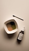 A neatly arranged scene featuring a small white bowl filled with four different colored powders, a small metallic spoon, and a brown glass bottle with a dropper labeled 'The Ordinary'. The bowl and bottle are placed on a light-colored surface, capturing a minimalist, clean aesthetic.