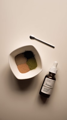 A neatly arranged scene featuring a small white bowl filled with four different colored powders, a small metallic spoon, and a brown glass bottle with a dropper labeled 'The Ordinary'. The bowl and bottle are placed on a light-colored surface, capturing a minimalist, clean aesthetic.