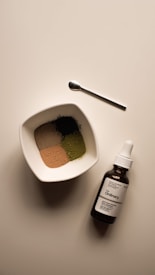 A neatly arranged scene featuring a small white bowl filled with four different colored powders, a small metallic spoon, and a brown glass bottle with a dropper labeled 'The Ordinary'. The bowl and bottle are placed on a light-colored surface, capturing a minimalist, clean aesthetic.