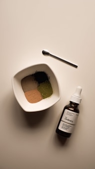 A neatly arranged scene featuring a small white bowl filled with four different colored powders, a small metallic spoon, and a brown glass bottle with a dropper labeled 'The Ordinary'. The bowl and bottle are placed on a light-colored surface, capturing a minimalist, clean aesthetic.