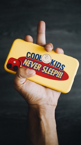 A hand is holding a yellow phone case with a design that includes a cartoon character and the text 'COOL KIDS NEVER SLEEP!!!'. The phone has a red back with visible camera lenses. The background is dark and out of focus.
