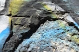 Rough textured wall surface with multicolored spray paint, showing vibrant hues including blue, yellow, and green across the cracked and uneven stone textures.