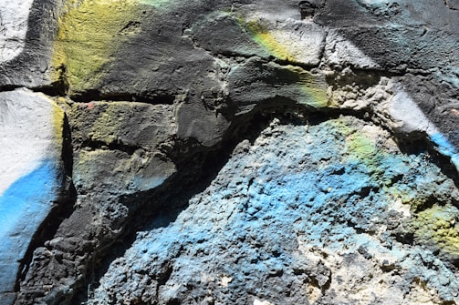 Rough textured wall surface with multicolored spray paint, showing vibrant hues including blue, yellow, and green across the cracked and uneven stone textures.
