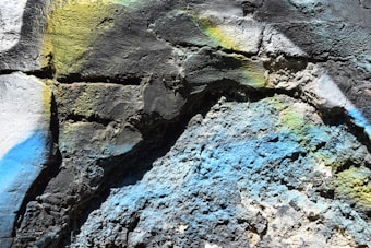 Rough textured wall surface with multicolored spray paint, showing vibrant hues including blue, yellow, and green across the cracked and uneven stone textures.