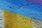 A textured wall surface painted with bold colors, including bright yellow, sky blue, and a hint of purple. The paint appears to be layered, creating distinct geometric patterns across the rough, concrete-like material.