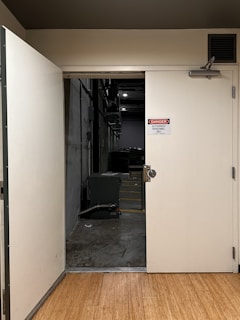An open door reveals an industrial or utility area with concrete flooring and walls. A sign on the door reads 'Danger Authorized Personnel Only'. Inside, there is a large green piece of equipment, some scattered debris on the floor, and dim overhead lighting.