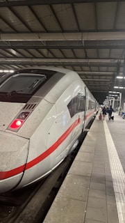 A sleek, modern white train with a red stripe, stationary at a well-lit indoor train station. The platform is tiled and partially visible, with a few travelers carrying luggage seen in the distance. The ceiling is high with metal beams and overhead lights.