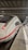 A sleek, modern white train with a red stripe, stationary at a well-lit indoor train station. The platform is tiled and partially visible, with a few travelers carrying luggage seen in the distance. The ceiling is high with metal beams and overhead lights.