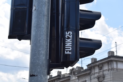 A close-up view of a traffic light mounted on a metal pole, with a backdrop of a partly cloudy sky and urban building rooftops. The word 'FUNK25' is visible on the side of the traffic light.