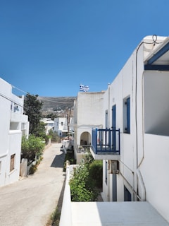 A narrow street flanked by whitewashed buildings with blue accents, typical of Mediterranean architecture. A Greek flag flutters on one of the buildings. The sky is clear and blue, and there is greenery lining the path.