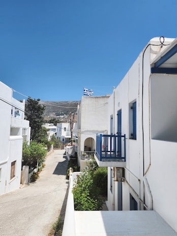 A narrow street flanked by whitewashed buildings with blue accents, typical of Mediterranean architecture. A Greek flag flutters on one of the buildings. The sky is clear and blue, and there is greenery lining the path.