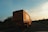 A large orange truck is driving on an open highway under a vast sky with scattered clouds. The surrounding landscape consists of greenery and trees, with some sections of the road visible in the distance. The lighting suggests early morning or late afternoon, casting a warm glow on the scene.