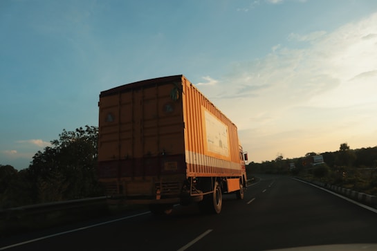A large orange truck is driving on an open highway under a vast sky with scattered clouds. The surrounding landscape consists of greenery and trees, with some sections of the road visible in the distance. The lighting suggests early morning or late afternoon, casting a warm glow on the scene.