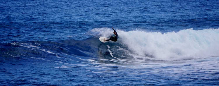 A surfer expertly rides a large, rolling wave in the ocean. The scene captures dynamic movement, with white water and spray surrounding the surfboard as the surfer balances with one arm raised.