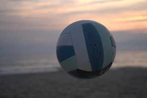 A volleyball is in mid-air against a beach backdrop with a soft, serene sunset. The sky is painted with hues of orange, pink, and gray, while the ocean and sand are subtly visible in the background, giving a tranquil feel.