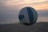 A volleyball is in mid-air against a beach backdrop with a soft, serene sunset. The sky is painted with hues of orange, pink, and gray, while the ocean and sand are subtly visible in the background, giving a tranquil feel.