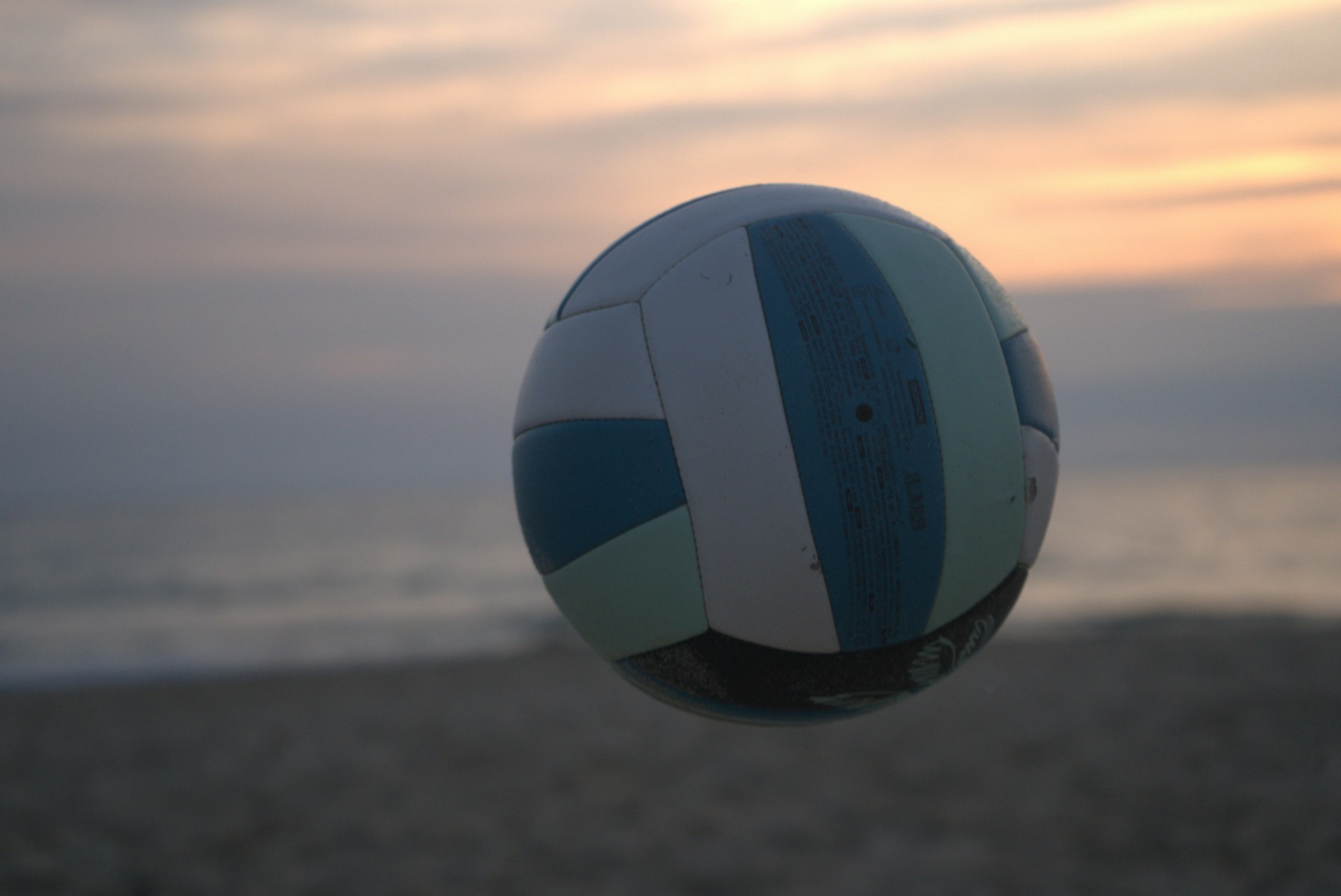 Dynamic beach volleyball scene with two players diving in the sand, clear blue sky and ocean in the background, dark gradient overlay.