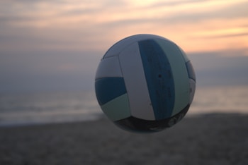 A volleyball is in mid-air against a beach backdrop with a soft, serene sunset. The sky is painted with hues of orange, pink, and gray, while the ocean and sand are subtly visible in the background, giving a tranquil feel.
