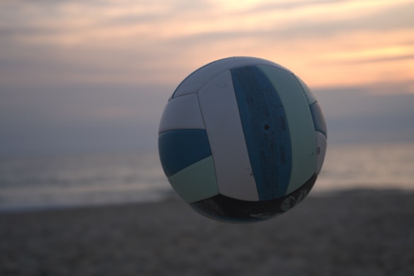 A volleyball is in mid-air against a beach backdrop with a soft, serene sunset. The sky is painted with hues of orange, pink, and gray, while the ocean and sand are subtly visible in the background, giving a tranquil feel.