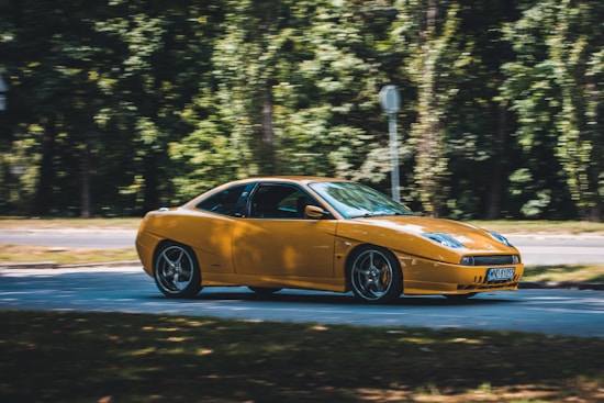 A sleek, yellow sports car is driving along a road surrounded by lush green trees. The car is in motion, with its blurred background indicating speed. Sunlight glints off its polished surface, highlighting its smooth lines and aerodynamic shape.