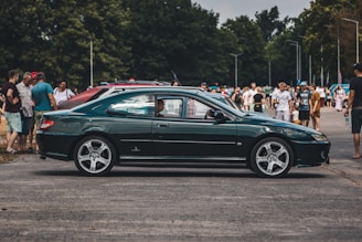 A dark green coupe car is parked outdoors with a group of people gathered around, suggesting a car show or event. Trees can be seen in the background, creating a relaxed, natural atmosphere. The car is well-polished, and the focus is on its sleek design and detailing.